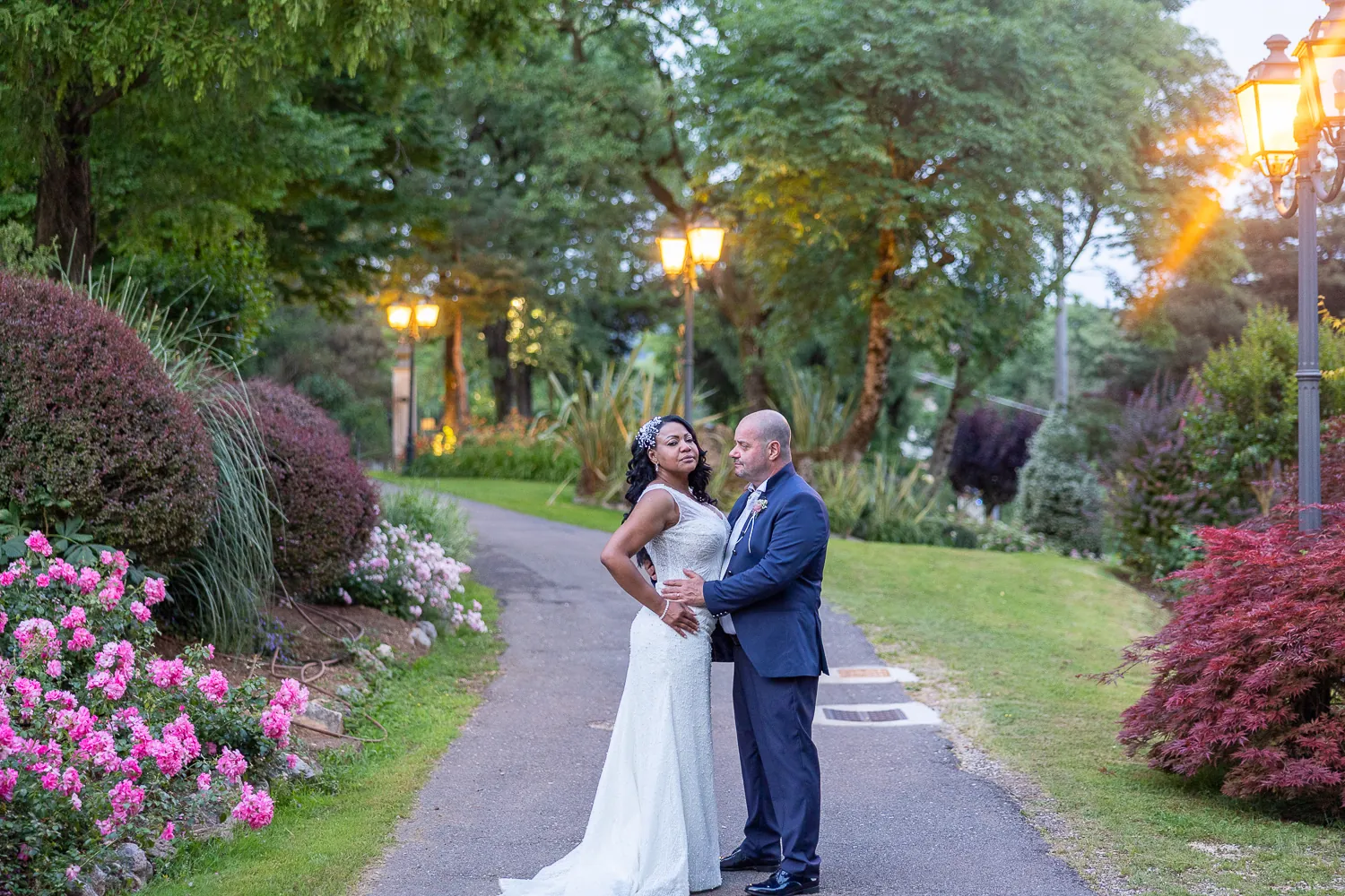 Ana e Andrea abbracciati sul viale del parco di Casa Mascagni al tramonto, tra cespugli di rose e lampioni accesi
