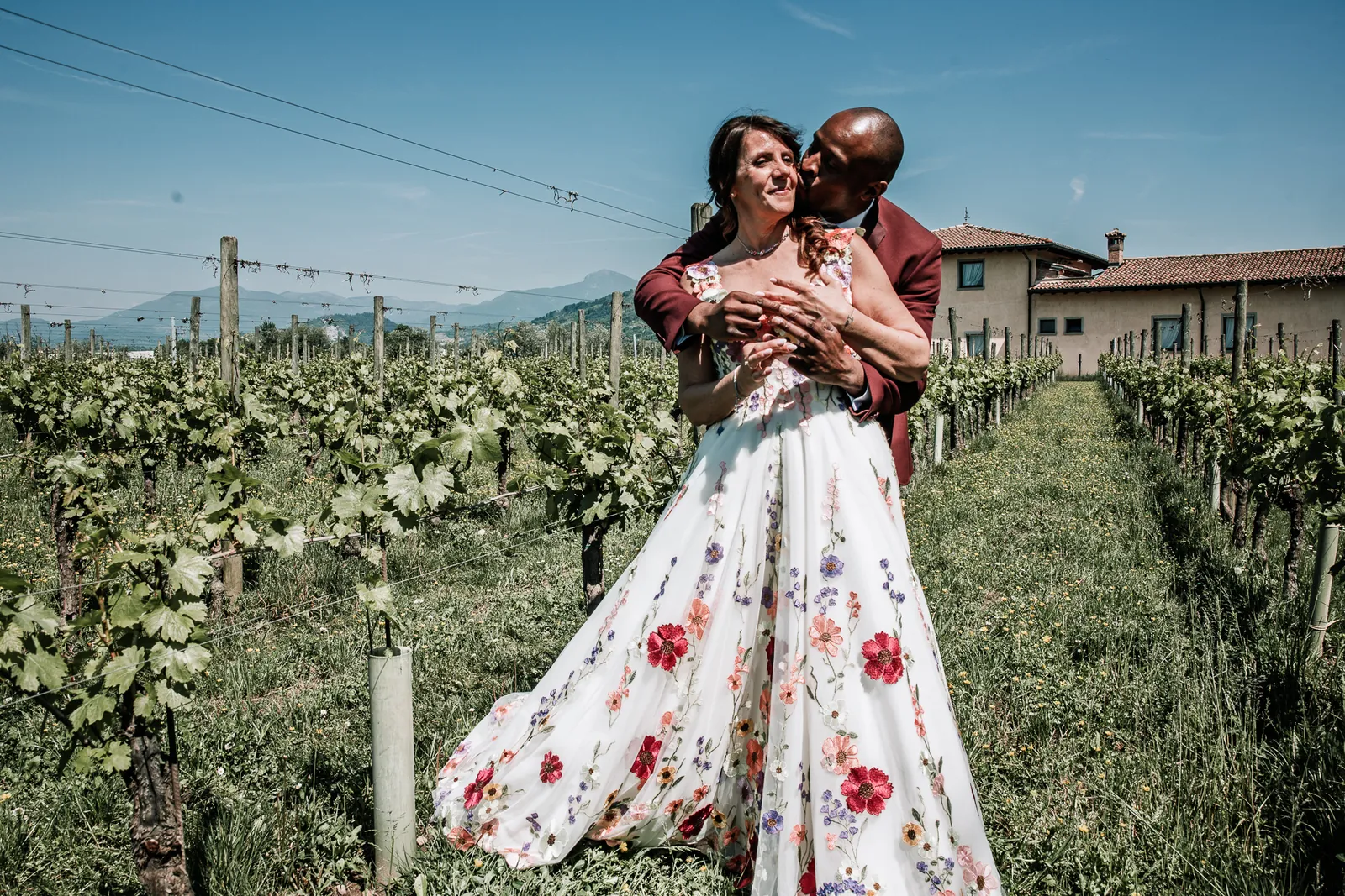 Katy e Dupe abbracciati tra i filari del vigneto di Cascina Carretto, con la cascina e le montagne sullo sfondo