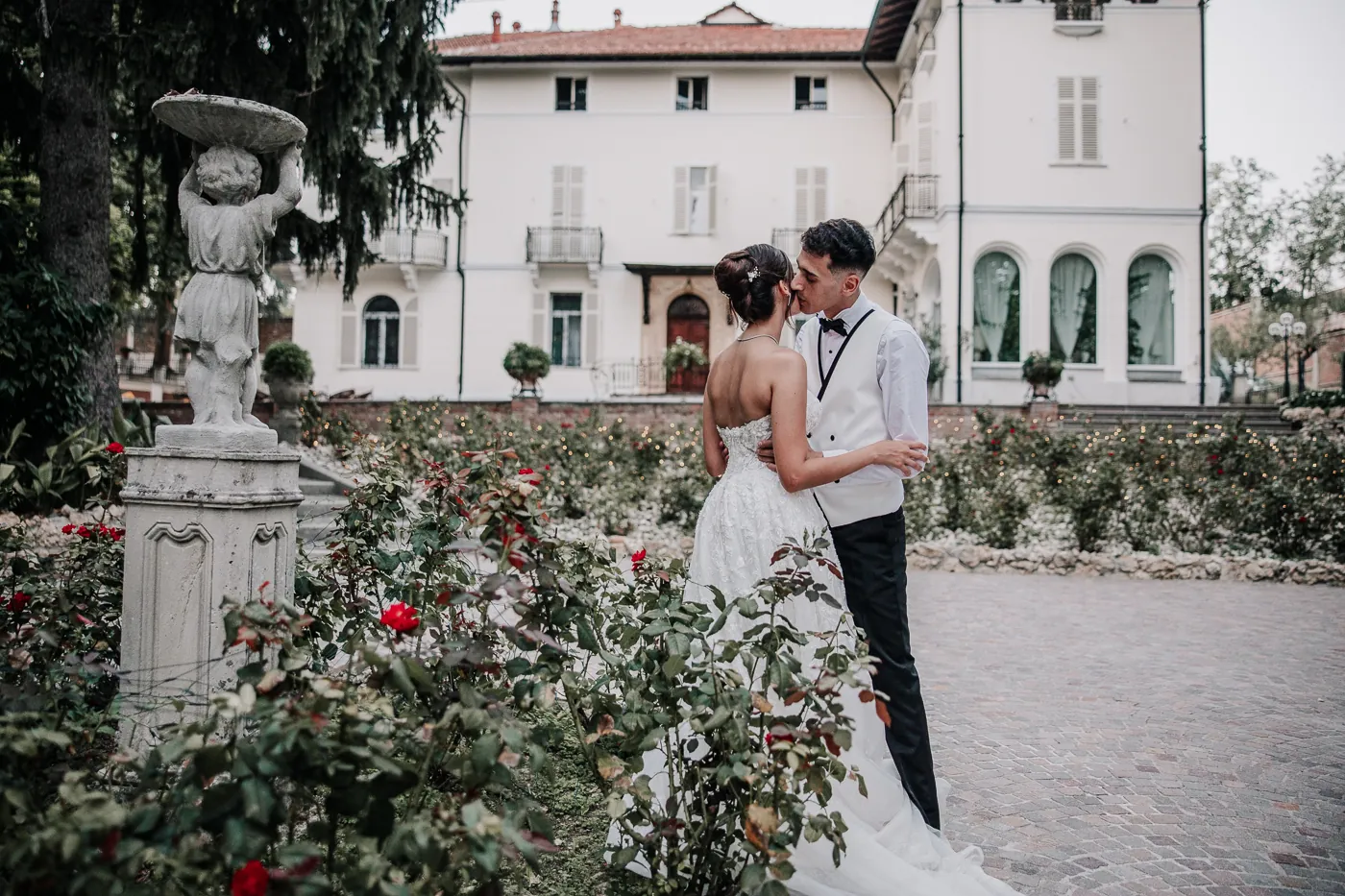 Maria e Mina si baciano nel roseto davanti alla villa bianca dei Giardini La Pergola, con una statua in primo piano