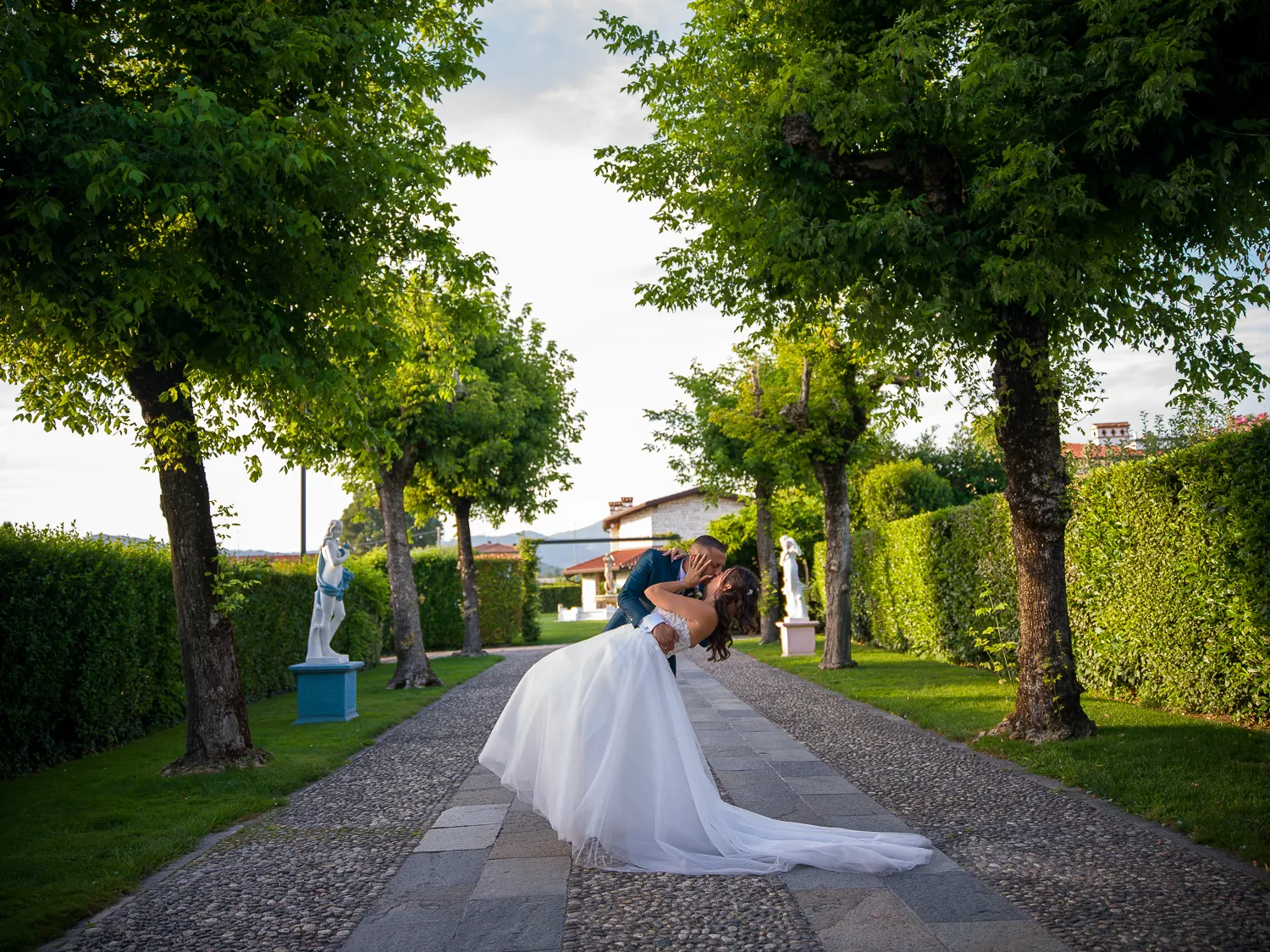 Alessia e Raffaele si baciano nel viale alberato di Villa Giardino, tra siepi e statue bianche, con il lungo strascico dell’abito sul selciato