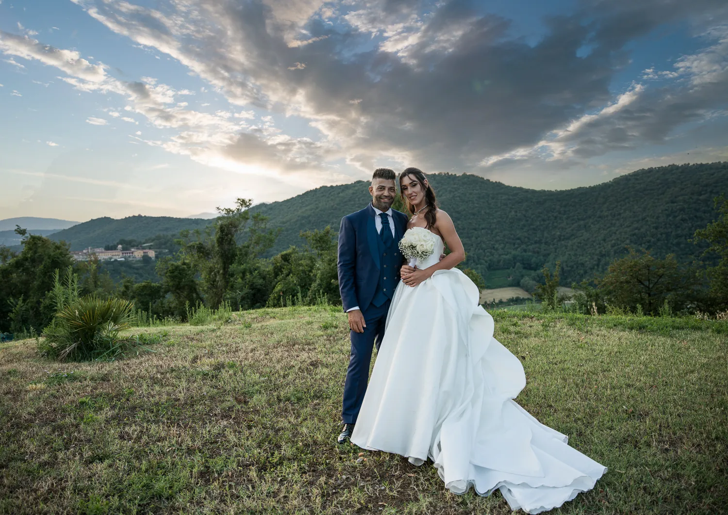 Axana e Mario sul prato in collina al tramonto, con i boschi della Franciacorta e un cielo di nuvole alle spalle