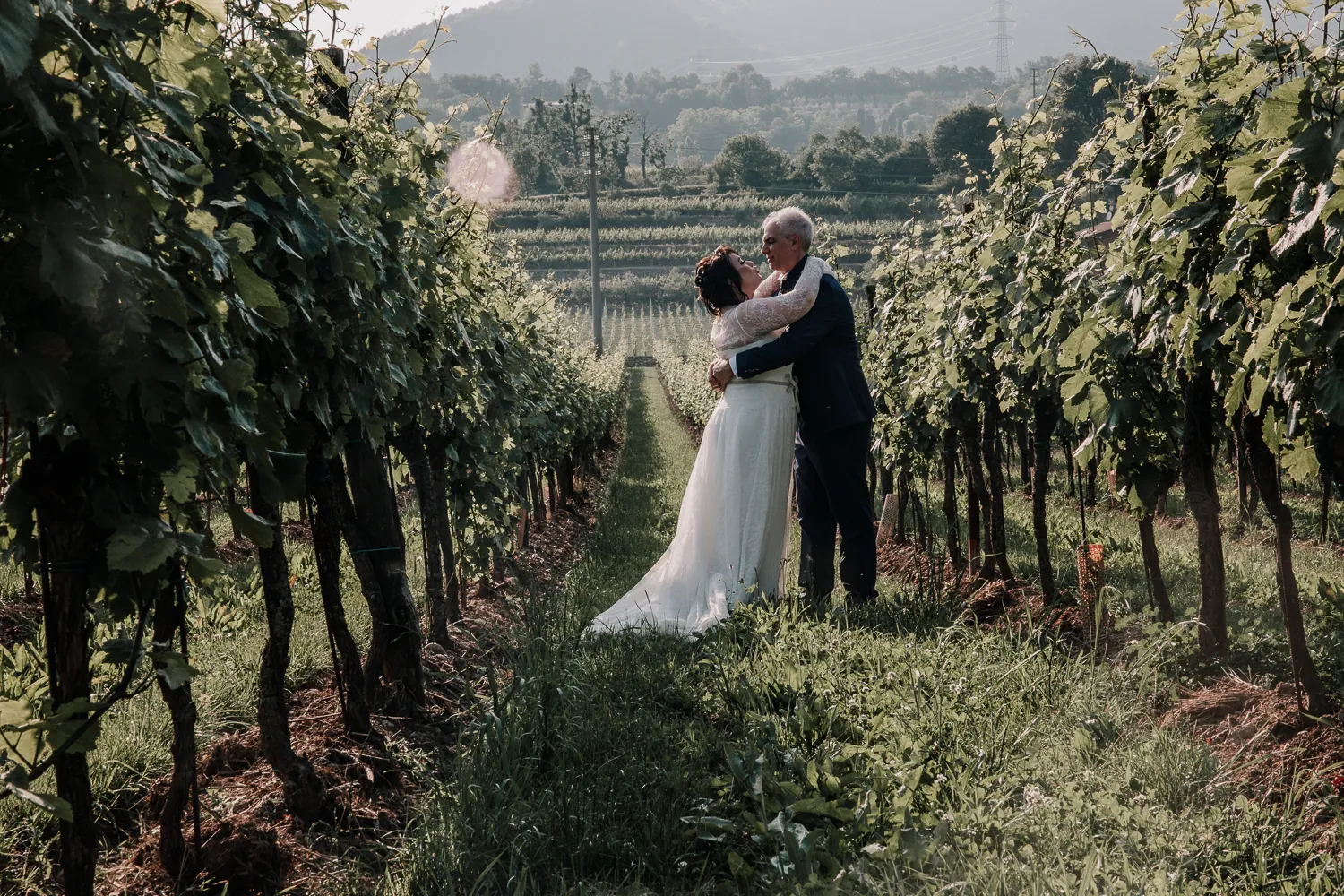 Ioana e Luca abbracciati tra i filari del vigneto in Franciacorta, con le colline sullo sfondo nella luce del tardo pomeriggio