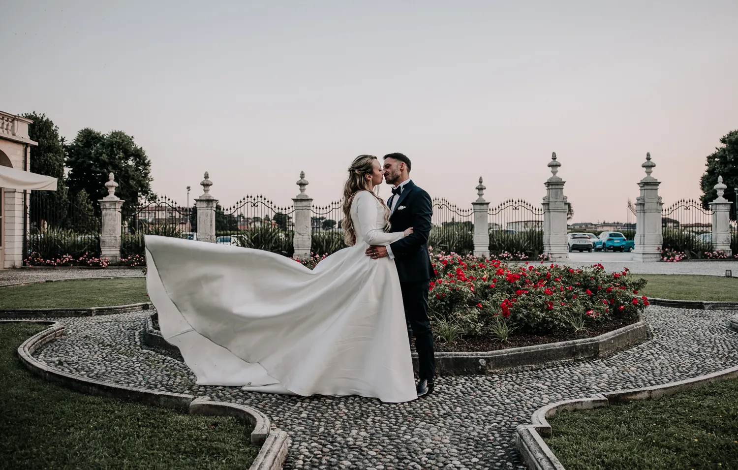 Luigi e Valentina si baciano sul vialetto acciottolato del giardino di Villa Fenaroli, con lo strascico dell’abito che vola, le aiuole di rose rosse e il cancello con i pilastri di pietra al tramonto